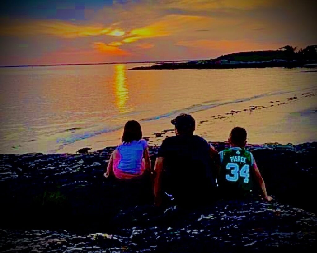 Three young people sitting together on a rocky shoreline overlooking the water